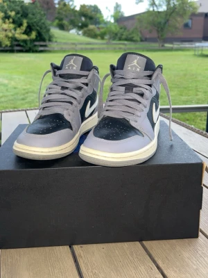 Nike Air Jordan 1 Mid grå/svart - Snygga Nike Air Jordan 1 Mid sneakers i grått och svart med vita detaljer och klassisk Swoosh på sidan. Skorna har en högre siluett, perforerad tåbox och gråa skosnören. Tillverkade i läder för en clean och stilren look.