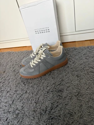 Maison marigela gats  - Snygga grå maison marigela gats sneakers med vita skosn och beige detaljer. Skorna har en klassisk låg siluett och en brun gummisula som ger en retrovibe. Ovandelen är i en mix av läder och mocka för en stilren look. Perfekta för dig som gillar minimalistisk design.