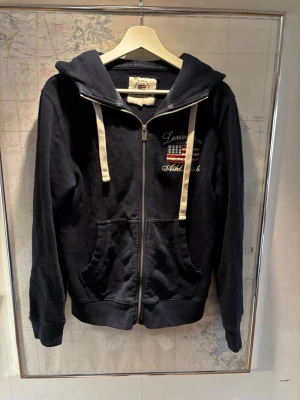 Zip hoodie från lexington - Snygg marinblå hoodie med dragkedja och vita snören. Har broderat tryck med amerikansk flagga och texten 'Lexington Athletics' på bröstet. Utrustad med kängurufickor och stor huva. Perfekt för en avslappnad och sportig stil.