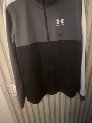 Svart och grå ziptröja Under Armour - Snygg ziptröja från Under Armour i svart och mörkgrått med vita detaljer på ärmarna. Tröjan har hel dragkedja framtill och logga på bröstet. Perfekt för träning eller chill. Tillverkad i mjukt funktionsmaterial.
