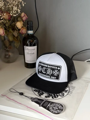 Svart Vit Chrome Hearts Keps - Snygg svart och vit truckerkeps med stort broderat patch framtill där det står 'Chrome Hearts Hollywood, USA' i vit text. Kepsen har mesh baktill och justerbar snapback. Perfekt för dig som gillar streetwear och vill sticka ut.