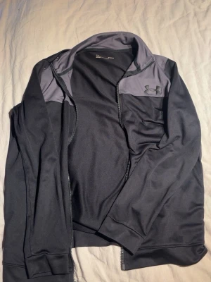 Under Armour zip hoodie - Svart zip hoodie från Under Armour med gråa axelpartier och diskret logga på bröstet. Tillverkad i ett mjukt, stretchigt funktionsmaterial som andas. Perfekt för träning eller chill. Klassisk passform med ribbade muddar och dragkedja framtill.
