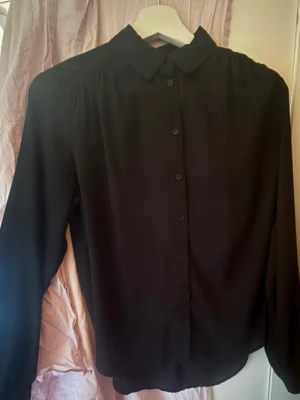 Svart klassisk skjortblus - Stilren svart skjortblus med knappar framtill och klassisk krage. Blusen har långärmad design och är tillverkad i ett tunt, mjukt tyg som ger ett snyggt fall. Perfekt att matcha med jeans eller kjol för en clean look.