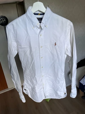 Vit skjorta från Polo Ralph Lauren - Klassisk vit skjorta från Polo Ralph Lauren med broderad logga på bröstet. Skjortan har knappar hela vägen, button-down krage och långa ärmar. Tillverkad i bomull för en skön och stilren look.