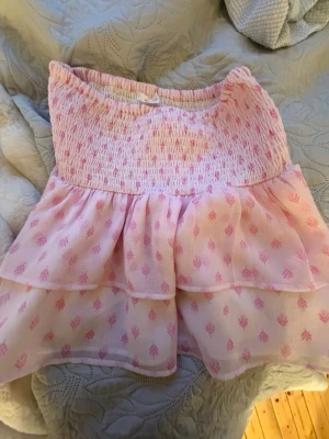 Söt rosa volangkjol med mönster - Supersöt kortkjol i ljusrosa med volanger och små rosa mönster. Kjolen har smock i midjan för skön passform och dubbla lager i tyget som ger en fluffig look. Perfekt för dig som gillar gulliga och feminina plagg.