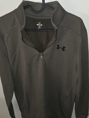Mörkgrön half-zip tröja Under Armour - Militärgrön Fleece-tröja från Under Armour. Tröjan är varm! Perfekt för lager-på-lager och sportiga outfits. Mycket bra skick!