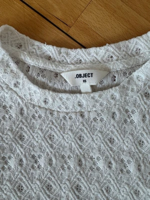Vit spetsblus från Object - Söt vit blus från Object i spets med puffiga korta ärmar och rund halsringning. Blusen har ett diskret mönster i tyget och känns både romantisk och trendig. Perfekt att styla med jeans eller kjol för en fräsch look.