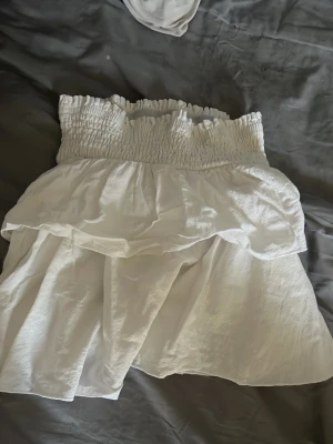 Vit kortkjol med volang - Söt vit kortkjol med smockad midja och volangdetalj. Kjolen har en luftig känsla och passar perfekt till varmare dagar. Enkel att matcha med olika toppar för en fräsch look.