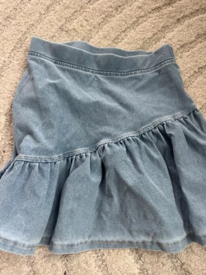 Ljusblå volangkjol i denim - Säljer en ljusblå jeanskjol med volang och asymmetrisk söm. Kjolen har en kort modell och är tillverkad i bomull. Perfekt för dig som gillar en avslappnad och trendig stil. Storlek 122/128