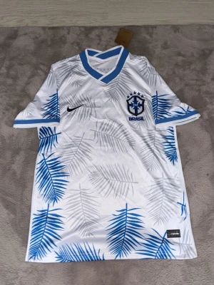 Vit Brasilien fotbollströja Nike - Snygg vit fotbollströja från Nike med Brasilien-logga på bröstet. Tröjan har blå detaljer vid krage och ärmslut samt blått palmbladsmönster över hela plagget. Tillverkad i ett lätt och ventilerande material, perfekt för träning eller match.