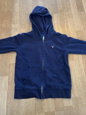 Mörkblå zip hoodie från Gant strl 11-12. 146-152 - Mörkblå hoodie med dragkedja från Gant. Klassisk design med huva, två stora fickor framtill och diskret Gant-logga på bröstet. Tillverkad i mjukt material som känns skönt mot huden. Perfekt för en avslappnad och stilren look.