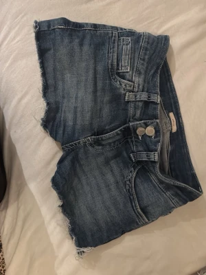 Blå jeansshorts med rå kant - Snygga blå jeansshorts med rå, fransig kant och dubbla knappar framtill. Shortsen har klassiska fickor och coola broderade detaljer på bakfickorna. Perfekta för varma dagar och ger en avslappnad vibe.