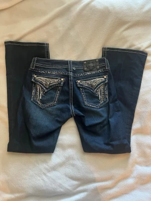 Mörkblå miss me jeans - Snygga mörkblå miss me jeans med utsvängda ben och vita kontrastsömmar. Baksidan har stora fickor dekorerade med silverfärgade nitar och strass i ett mönster. Modellen heter easy boot 🌟 storleken är 26 (ca storlek S), Innerbenslängd: 78cm 🔥 köpta secondhand men ändå bra skick!!