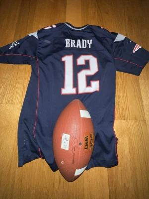Mörkblå NFL-tröja med nummer 12. Tom Brady tröja! - Säljer en mörkblå amerikansk fotbollströja med namnet BRADY och nummer 12 på ryggen. Tröjan har röda och vita detaljer samt grå axelpartier. Perfekt för dig som gillar amerikansk fotboll och vill ha en ikonisk supportertröja av Tom Brady. Pris kan diskuseras