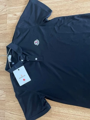 Svart pikétröja  - Klassisk svart pikétröja med korta ärmar och knappslå framtill. ikoniska logga broderad på bröstet och är tillverkad i mjuk bomull. Perfekt till både jeans och shorts. Aldrig använd det är storlek s på tröjan men den passar som m 