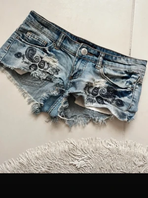 Blå slitna jeansshorts med brodyr - Säljer ett par ljusblå jeansshorts med slitna kanter och råa avslut. Shortsen har snygga broderade detaljer i svart på framfickorna och en liten brodyr på ena bakfickan. Klassisk femficksmodell med knapp och dragkedja. Perfekta för sommaren! passar xxs och xs