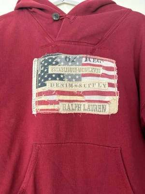 Vinröd hoodie från Ralph Lauren - Vinröd hoodie med stor magficka och huva med knappdetalj. På bröstet finns en stor patch med amerikansk flagga och texten 'Denim & Supply Ralph Lauren'. Perfekt för en avslappnad och cool stil. Något stor i storleken, passar nog M-L