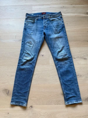 Slim Jeans med slitningar revor - 32/34. En söm och en fläck, syns på sista bilden. Säljer ett par blå slim jeans med slitningar och hål på båda benen. Jeansen har klassisk femficksmodell. Materialet är denim i bomull med stretch för extra komfort. Perfekta för en avslappnad och trendig look. 