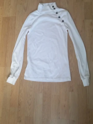 Vit polotopp med knappar - Snygg vit polotopp med lång ärm och dekorativa knappar längs axeln. Toppen har en hög krage och är figurnära i modellen, perfekt för att skapa en clean och stilren look. Osäker på storlek men tror att den är S-M.