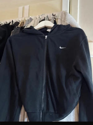 Svart zip hoodie från Nike - Säljer en svart hoodie från Nike med dragkedja och huva. Klassisk design med liten vit Nike-logga på bröstet. Tillverkad i mjukt material som passar perfekt till chill eller träning. Snygg och enkel att matcha med allt.