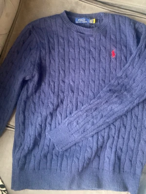 Polo Ralph lauren sweatshirt  - Snygg lila kabelstickad tröja från Polo Ralph Lauren med rund halsringning och klassisk röd logga broderad på bröstet. Tröjan har långa ärmar och ribbade muddar vid ärmslut och nederkant. Perfekt för dig som gillar stilrena stickade plagg.
