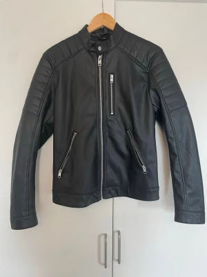 Svart biker skinnjacka med dragkedjor - Säljer en svart bikerinspirerad skinnjacka med ståkrage och tryckknapp. Jackan har tre dragkedjeförsedda fickor framtill och vadderade detaljer på axlar och ärmar för en cool look. Materialet är slätt och ger en klassisk rockig känsla.