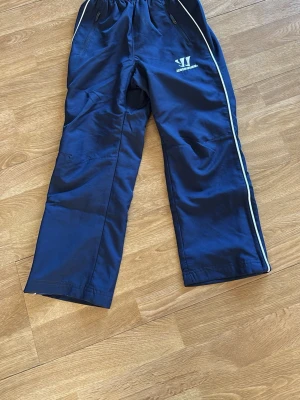 Blå hockeybyxor från Warrior - Säljer ett par blå hockeybyxor som är lätt fodrade från Warrior med vita detaljer längs sidorna och resår i midjan. Byxorna har fickor med dragkedja och är perfekta för sport eller träning. Materialet är lätt och bekvämt för aktiva dagar. Benskydd under får plats.
