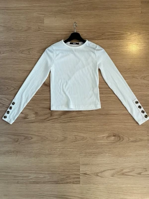 Vit ribbad långärmad topp - Säljer en vit ribbad långärmad topp med rund hals och dekorativa knappar i brunt på ärmsluten. Toppen har en rak passform och är tillverkad i ett stretchigt material som sitter snyggt på kroppen. Perfekt att styla med jeans eller kjol.