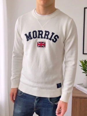Vit stickad tröja från Morris - Vit stickad tröja från Morris med mörkblå text och en broderad brittisk flagga på bröstet. Tröjan har rund halsringning, ribbade muddar och nederkant samt en liten patch nertill. Perfekt för en clean och klassisk stil.