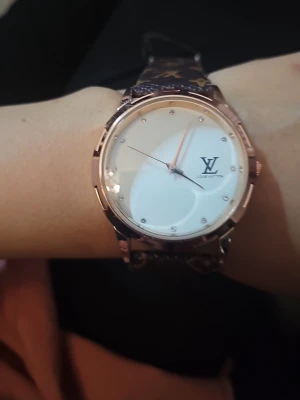  klocka med monogram - Stilren klocka med rund vit urtavla och guldfärgat boett. Armbandet är i brunt läder med klassiskt LV-monogram i guld. Urtavlan har minimalistiska markeringar och LV-logga. Perfekt accessoar för dig som gillar lyxig och tidlös design.