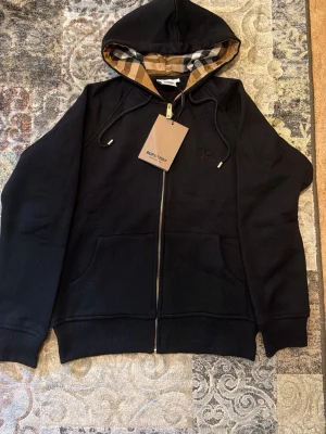 Svart Burberry kofta med beige huva - Jag säljer varan för att den inte passar mig.                                                                       Tröjan är helt ny och oanvänd och passar perfekt till hösten och kalla sommarkvällar.