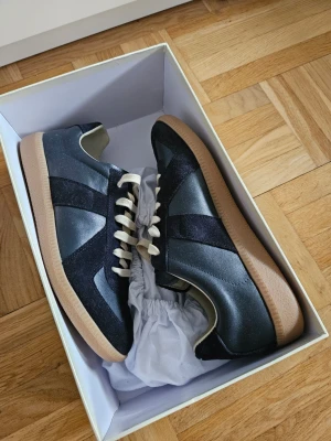 Maison Margiela Navy Gats - Stilrena Maison Margiela Paris Navy GAT sneakers med kontrasterande mockadetaljer och beige snörning. Skorna har en klassisk låg siluett och en ljusbrun gummisula som ger en retrovibe. Materialmix av läder och mocka gör dem både snygga och bekväma. Perfekta för dig som gillar att stå ut men inte vara för flashy.