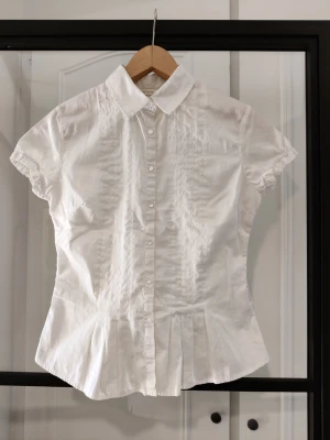 Vintage Vit kortärmad blus med knappar - Stilren vit blus med korta ärmar och klassisk krage. Blusen har knäppning framtill och dekorativa veck nedtill som ger en snygg siluett. Tillverkad i ett lätt tyg som känns fräscht och passar till många olika looks. Vintage shirt from the 90s, centered at the waist  Super flattering, trust me the pictures don't give it justice  Tight fit best for smaller chest  In great condition, no damages  Don't hesitate to make an offer. I invite you to look at my other listings and to make a bundle 🫶