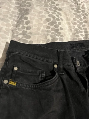 Svarta raka jeans tiger of Sweden  - Säljer ett par svarta jeans med klassisk femficksmodell och raka ben. Jeansen har silverfärgade nitar och knapp, samt en liten gul detalj vid myntfickan. Materialet är denim i bomull och passformen är normal.