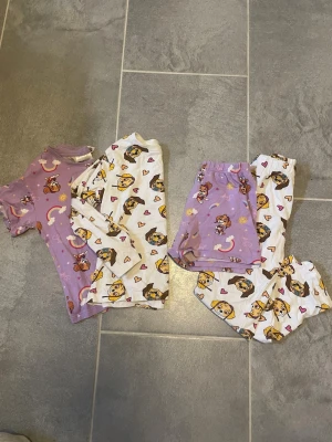 2-pack Söta Paw Patrol pyjamas-set - Två pyjamas-set med Paw Patrol-motiv. Ett set är lila med kortärmad tröja och shorts, det andra är vitt med långärmad tröja och byxor. Båda har tryck av Skye och små hjärtan. Perfekta för mysiga kvällar och sömn. Mjukt och bekvämt material.