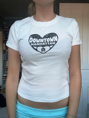 Vit t-shirt med Downtown Manhattan-tryck - Vit t-shirt med svart tryck i form av ett hjärta och texten 'Downtown Manhattan' på bröstet. Modellen är figurnära med korta ärmar och rund hals. Perfekt för en avslappnad och trendig stil.