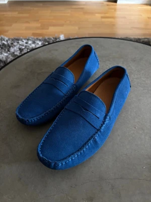 Blå mocka loafers herr - Snygga loafers i klarblå mocka med klassisk pennyloafers-design. Skorna har rundad tå, dekorativ söm runt ovandelen och gummidetaljer på hälen. Aldrig använda då dem är för små, köpta i marbella😂