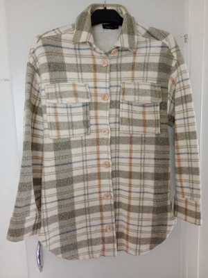 Rutig overshirt i beige och grönt - Snygg overshirt med rutigt mönster i beige, grönt, vitt och inslag av blått och orange. Två bröstfickor med lock, stora knappar framtill och klassisk krage. Tillverkad i mjukt, tjockt tyg som ger en avslappnad och trendig look.