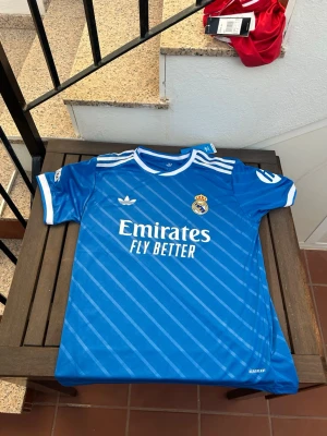 Blå Real Madrid matchtröja Adidas - Snygg blå Real Madrid matchtröja från Adidas med vita detaljer och diagonala ränder. Tröjan har klubbmärke på bröstet, Adidas-logga och sponsortryck 'Emirates Fly Better' framtill. Tillverkad i lätt och ventilerande material, perfekt för fotbollsträning eller match.