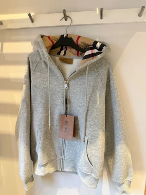 Grå zip hoodie från Burberry - Snygg grå zip hoodie från Burberry med klassiskt rutigt foder i huvan. Hoodien har dragsko i huvan, fickor fram och ribbade muddar. Perfekt för dig som vill ha en stilren men ändå lyxig look. Materialet är mjukt och bekvämt, troligen bomullsblandning. Storleken är M men den känns som s