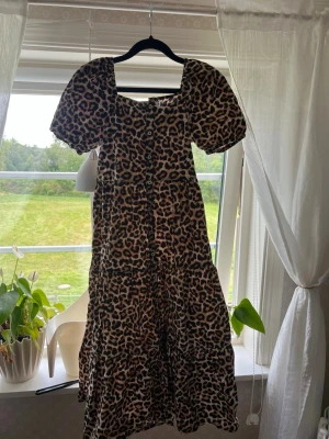 Lång leopardklänning med puffärm - Snygg långklänning med leopardmönster och puffiga kortärmar. Klänningen har knappar framtill och en luftig passform som ger en cool och trendig look. Perfekt för dig som vill sticka ut med ett djurmönster.