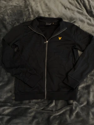 Svart ziptröja från Lyle & Scott - Snygg svart ziptröja från Lyle & Scott med gul broderad logga på bröstet. Tröjan har hög krage, långa ärmar och ribbade muddar. Tillverkad i mjukt material och har två fickor framtill. Perfekt för en avslappnad och stilren look.