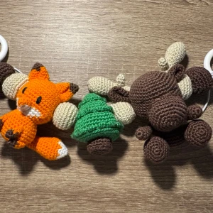 Virkad barnvagnshänge med djur - Säljer ett handgjort barnvagnshänge med virkade figurer: en orange räv, en grön gran och en brun älg. Alla figurer är gjorda i bomullsgarn och sitter fast på ett vitt band med ringar i plast. Perfekt som söt dekoration till barnvagnen.