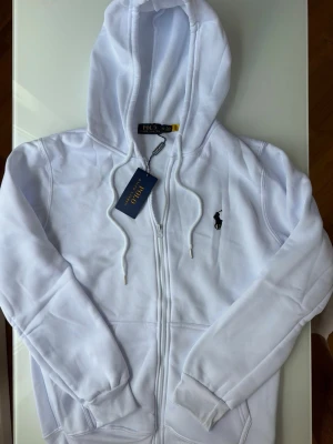 Vit hoodie från Polo Ralph Lauren - Snygg vit hoodie från Polo Ralph Lauren med dragkedja framtill och klassisk huva med dragsko. Ikoniska svarta loggan broderad på bröstet. Tillverkad i mjukt material och har ribbade muddar vid ärmar och nederkant. Perfekt för en clean och stilren look.