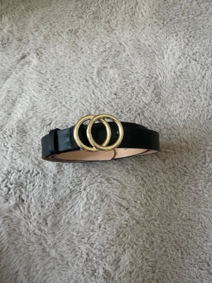 Svart bälte med guldfärgat spänne - Stilrent svart bälte i skinnimitation med ett snyggt guldfärgat spänne i form av två ringar. Perfekt accessoar för att lyfta vilken outfit som helst. Justerbar längd med flera hål för bästa passform.