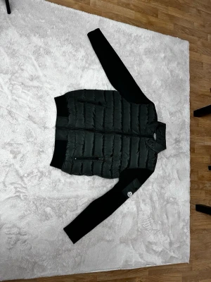 Moncler Cardigan kofta - Snygg svart pufferjacka från Moncler med quiltad kropp i glansigt material och svarta stickade ärmar. Jackan har hög krage, dragkedja framtill och två fickor med dragkedja. Moncler-logga på ena ärmen ger extra stilpoäng.