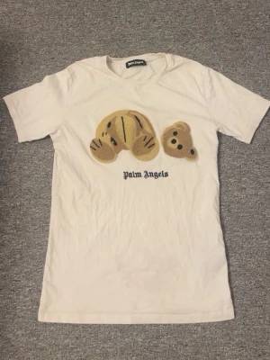 Vit Palm Angels t-shirt med björn - Vit t-shirt från Palm Angels med korta ärmar och ett stort broderat motiv av en nallebjörn på framsidan. T-shirten har rund hals och texten 'Palm Angels' under motivet. Perfekt för dig som gillar streetwear och unika detaljer.