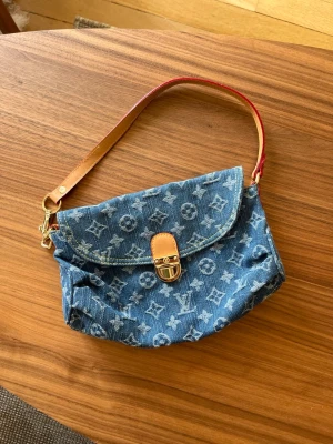 Blå denim handväska med monogram - Trendig handväska i blå denim med ikoniskt monogrammönster och detaljer i brunt läder. Väskan har ett lock med guldfärgat spänne och en kort axelrem i läder. Perfekt för dig som gillar en cool och avslappnad stil.