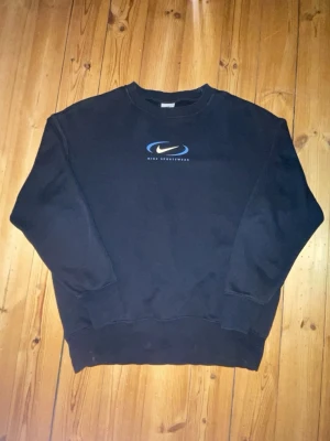 Svart Nike sweatshirt med blå logga - Säljer en svart sweatshirt från Nike med blå och vit logga samt texten 'Nike Sportswear' på bröstet. Tröjan har rund halsringning, ribbade muddar vid ärmslut och nederkant. Perfekt för en avslappnad och sportig stil.