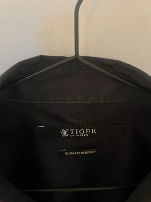 Svart skjorta från Tiger of Sweden - Stilren svart skjorta från Tiger of Sweden med slim fit stretch-passform. Klassisk krage och långa ärmar, perfekt för dig som gillar en smalare siluett. Skjortan har knappar framtill och är tillverkad i ett mjukt, stretchigt material.